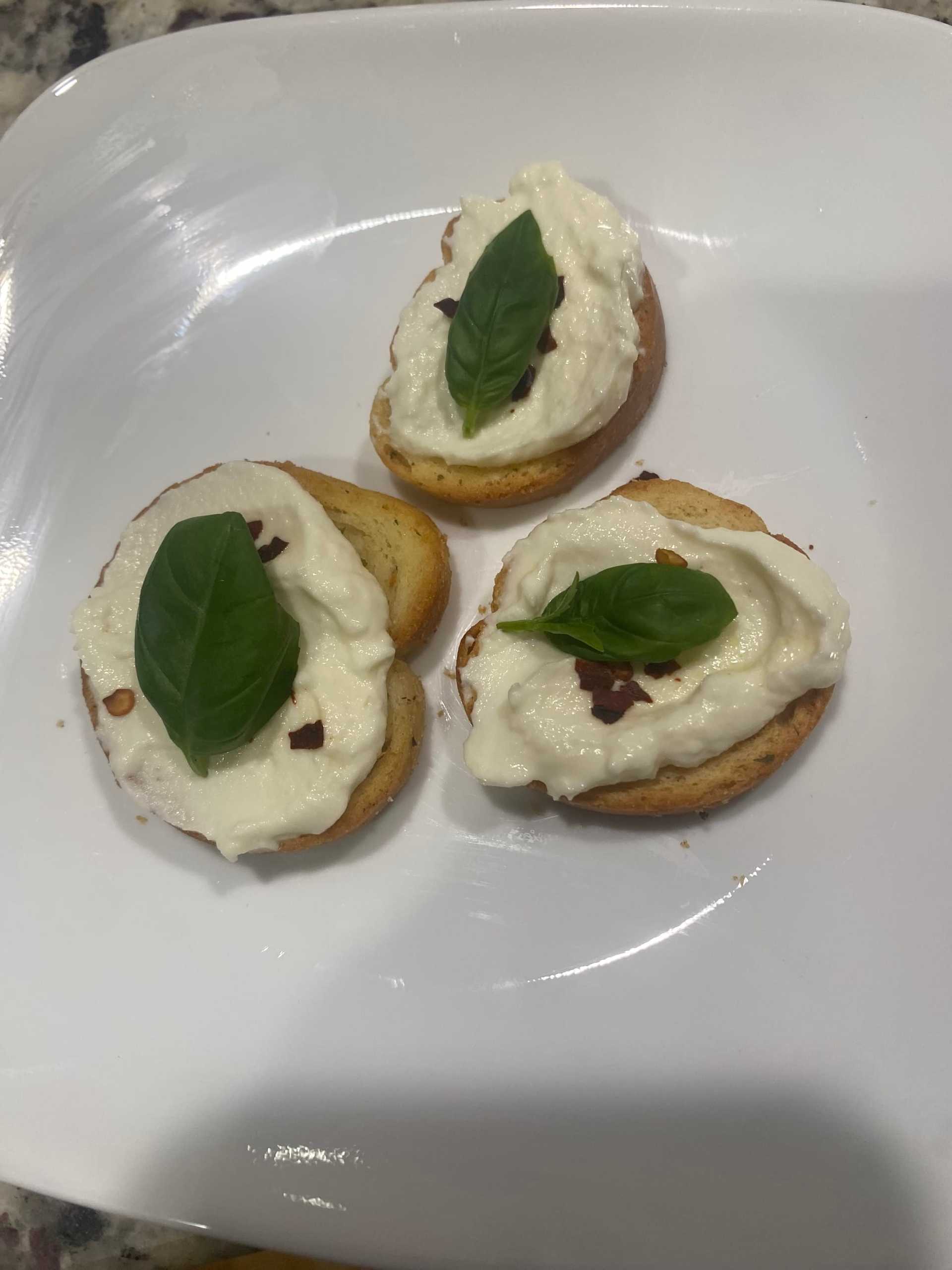 Bruschetta with ricotta, basil, and red pepper flakes on a white plate.